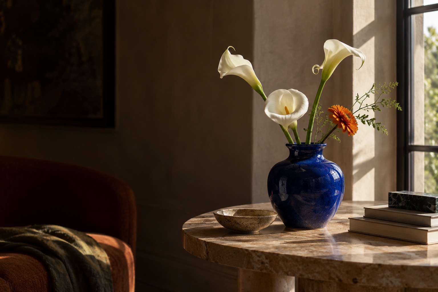 White calla lilies and an orange flower in a blue vase by a sunlit window